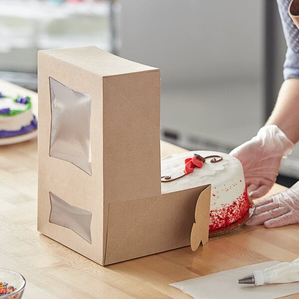 A woman wearing white gloves cuts a cake in a Baker's Mark Kraft bakery box with a window.
