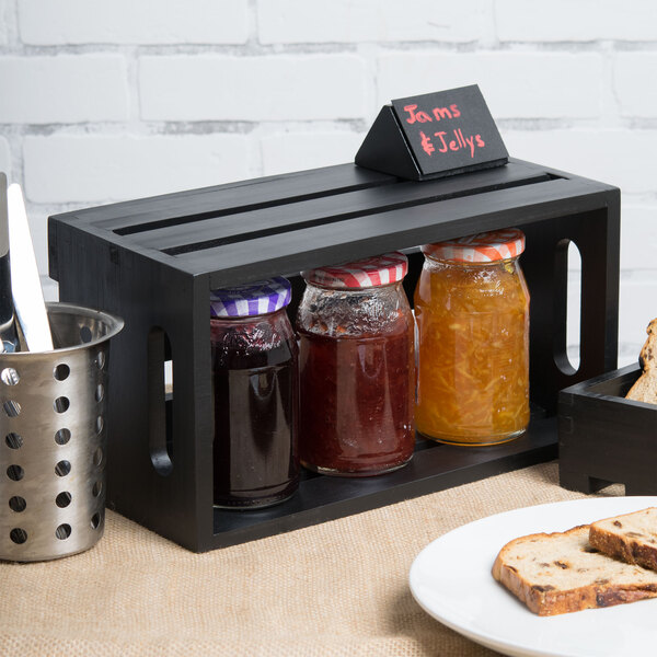 A black wooden American Metalcraft crate holding jars of jam and a plate of bread on a table.