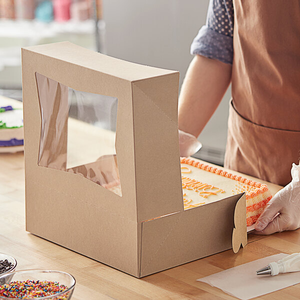 A person in a brown apron and gloves opening a Baker's Mark Kraft bakery box with a window.