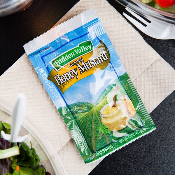 A plate with a salad topped with a Hidden Valley Golden Honey Mustard dressing packet on a table.