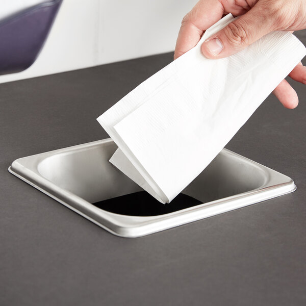 A hand dropping a white napkin into a rectangular stainless steel in-counter trash chute.