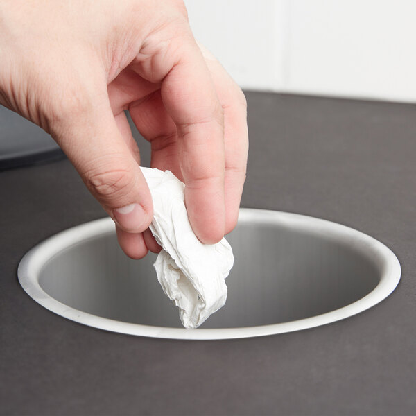 A hand dropping a tissue into a round stainless steel trash chute in a counter.
