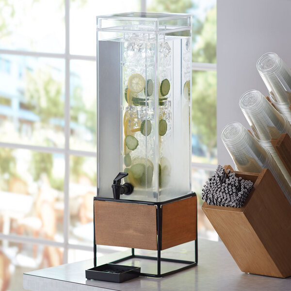 A Cal-Mil Madera beverage dispenser with a wooden and metal base and an infusion chamber filled with water.