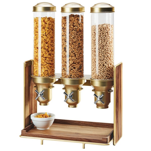 A Cal-Mil walnut and brass triple canister cereal dispenser on a hotel buffet counter with cereal in the clear glass containers.