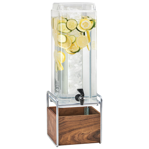 A Cal-Mil square beverage dispenser filled with water, lemons, and cucumbers on a walnut and chrome base.