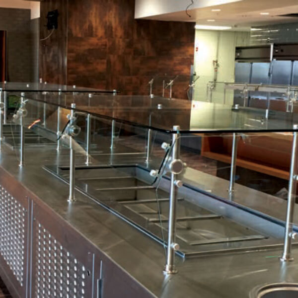 An Advance Tabco double sided self service food shield with stainless steel shelves on a large glass counter.