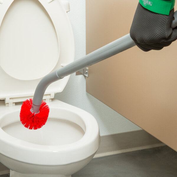 A hand holding a Unger Ergo toilet bowl brush with a red brush head cleaning a toilet.