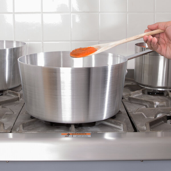 A person stirring a silver Vollrath aluminum sauce pan with a wooden spoon.