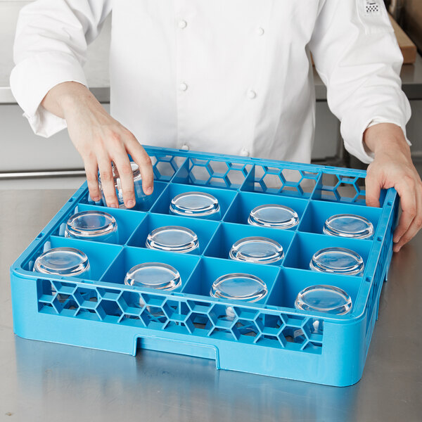 A person wearing a white coat and holding a blue Carlisle glass rack with clear glasses inside.