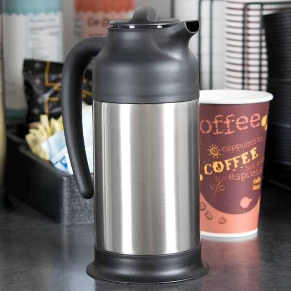 A Choice stainless steel coffee carafe on a counter next to a cup of coffee.