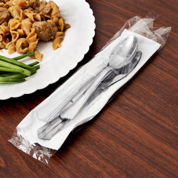 A table with a plate of pasta, vegetables, and a wrapped stainless steel look plastic fork and spoon.
