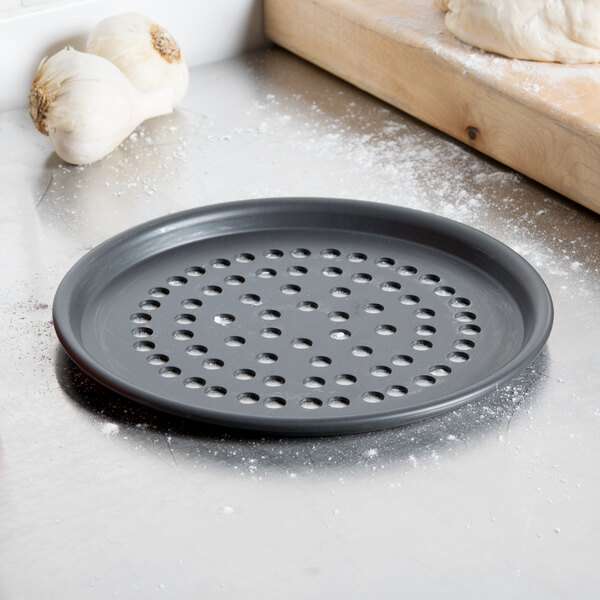 A black American Metalcraft Super Perforated Pizza Pan with garlic on a wooden cutting board.