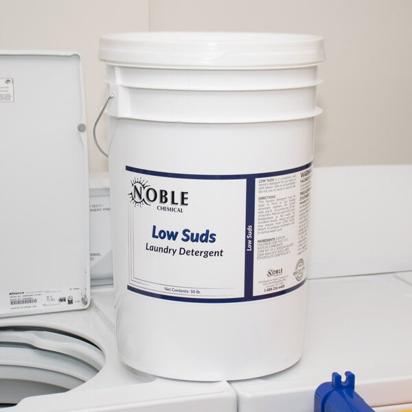 A white bucket of Noble Chemical Low Suds Concentrated Laundry Detergent sitting on a counter next to a washing machine.