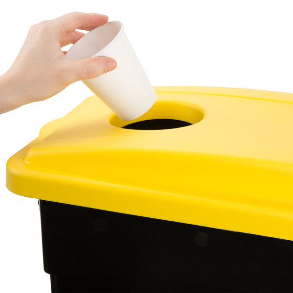 A hand putting a white cup into a yellow and black rectangular trash can lid.