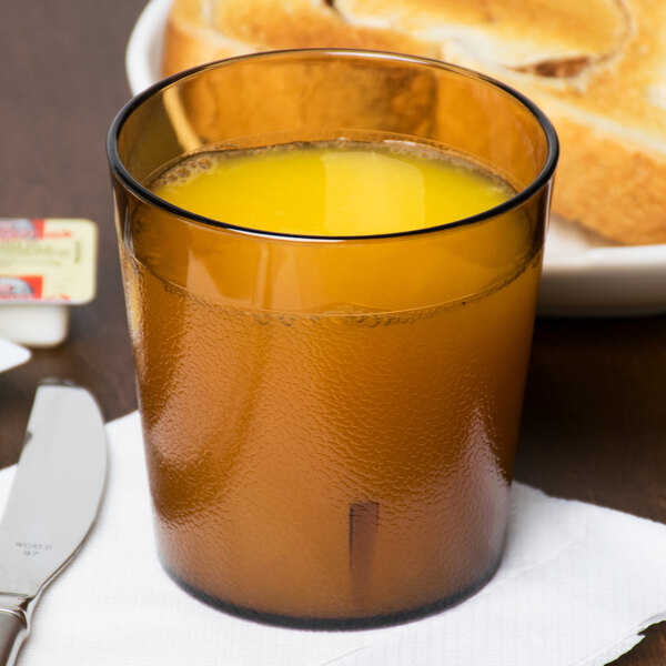 A Cambro amber plastic tumbler filled with orange juice on a table with a plate of bread and a knife.