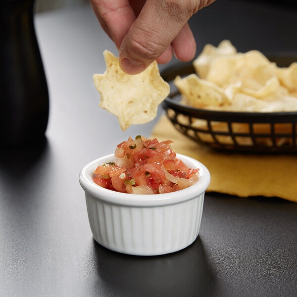 A person holding a Tuxton white fluted ramekin filled with salsa and dipping a chip.