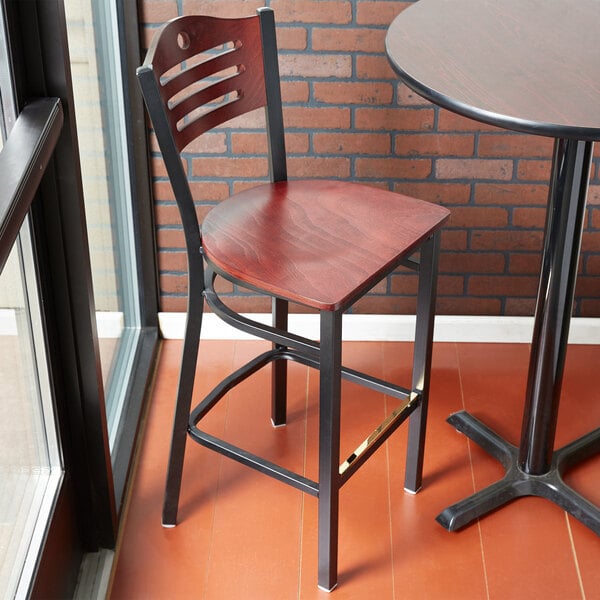 A Lancaster Table & Seating black bistro bar stool with mahogany wood seat and back on a table in front of a brick wall.