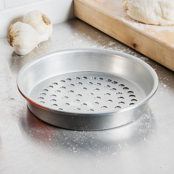 An American Metalcraft tin-plated steel pizza pan with holes on a counter next to a wooden cutting board and garlic.