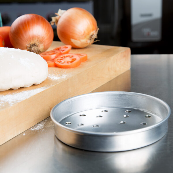 An American Metalcraft aluminum pizza pan with holes on a counter next to tomatoes.