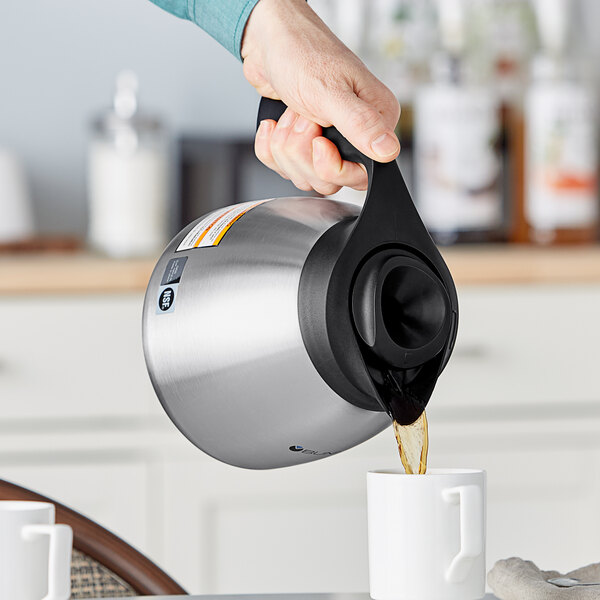 A person using a Bunn stainless steel coffee carafe with a black top to pour coffee into a white mug.