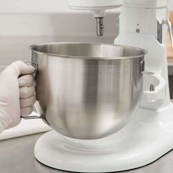 A person in white gloves holding a silver KitchenAid mixing bowl.