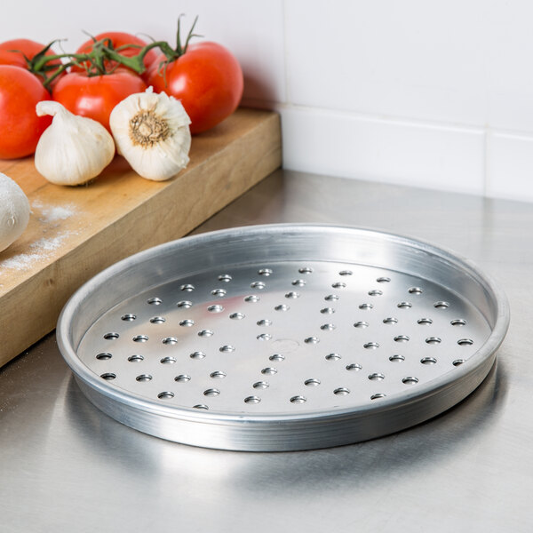 An American Metalcraft heavy weight aluminum pizza pan with holes next to tomatoes and garlic on a cutting board.
