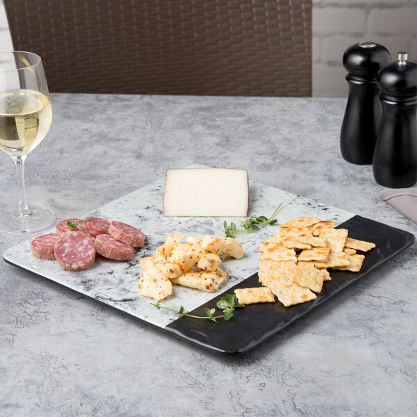 A white marble square serving board with a black border holding a plate of crackers, cheese, and wine.