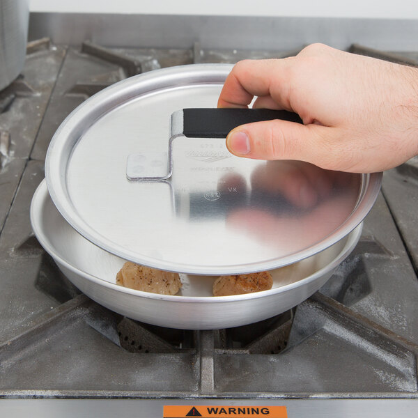 A hand holding a Vollrath Wear-Ever metal lid over a pan on a stove.