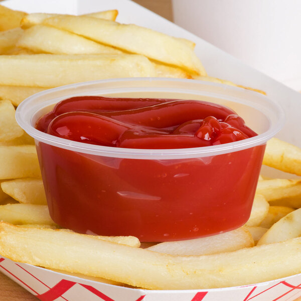 A clear oval plastic souffle cup filled with ketchup on a container of french fries.