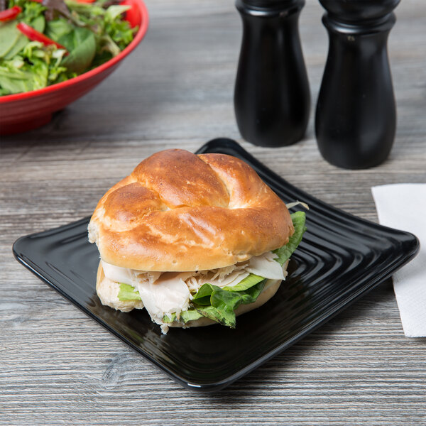 A Milano black melamine square plate with a sandwich on it and a bowl of salad on the counter.