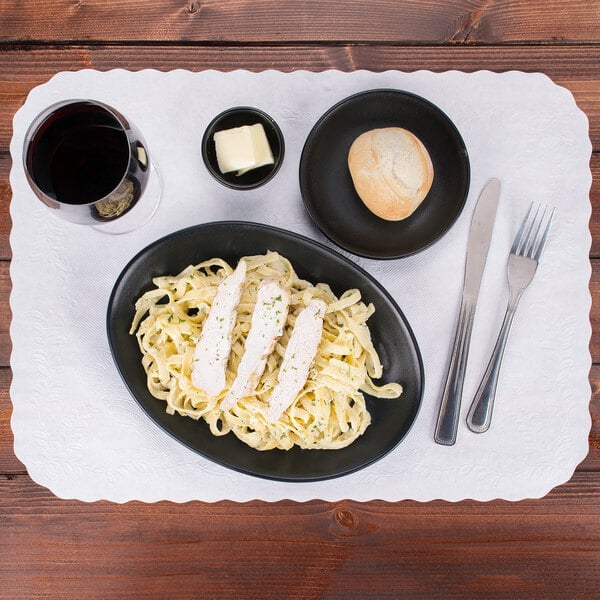 A white disposable traymat with a scalloped edge on a table set with a plate of pasta, bread, and a glass of wine.