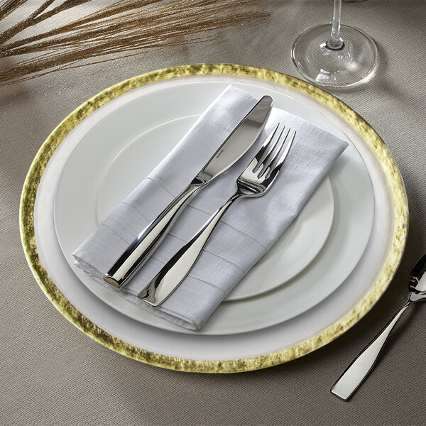 A white charger plate with a hammered gold rim on a white background with silverware and a white napkin.