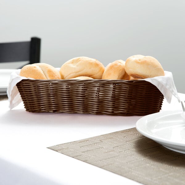 A Tablecraft brown rattan bread basket with rolls on a table.