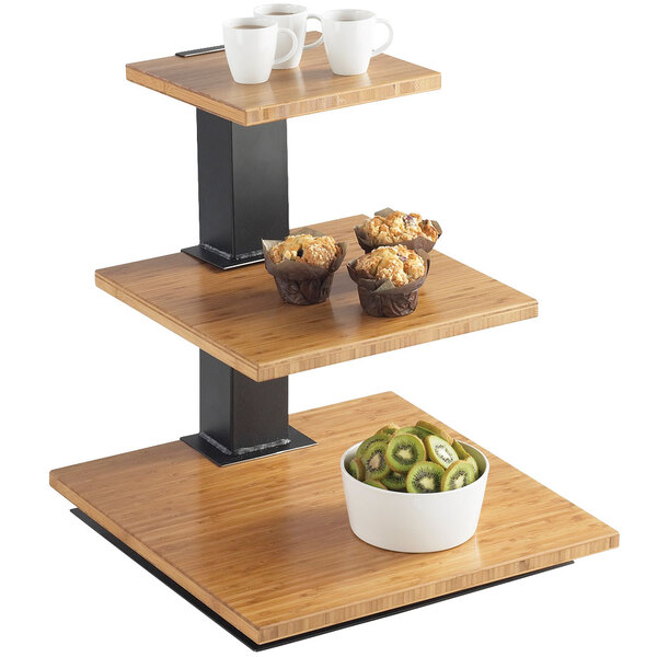 A Cal-Mil bamboo 3 tier riser with bamboo shelves holding bowls, cups, and fruit on a wooden table.