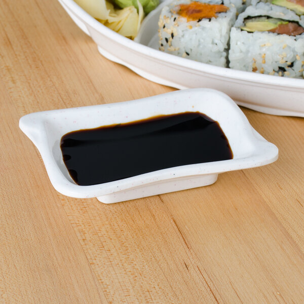 A white Thunder Group Blue Bamboo wave sauce dish with black liquid on a table with sushi rolls.