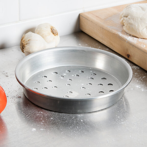 An American Metalcraft tin-plated steel pizza pan with holes on a counter next to garlic and a cutting board.