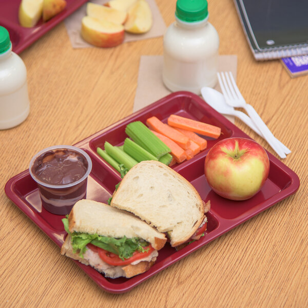 A Carlisle dark cranberry melamine 4 compartment tray with a sandwich, apple, vegetables, and chocolate pudding on a table.