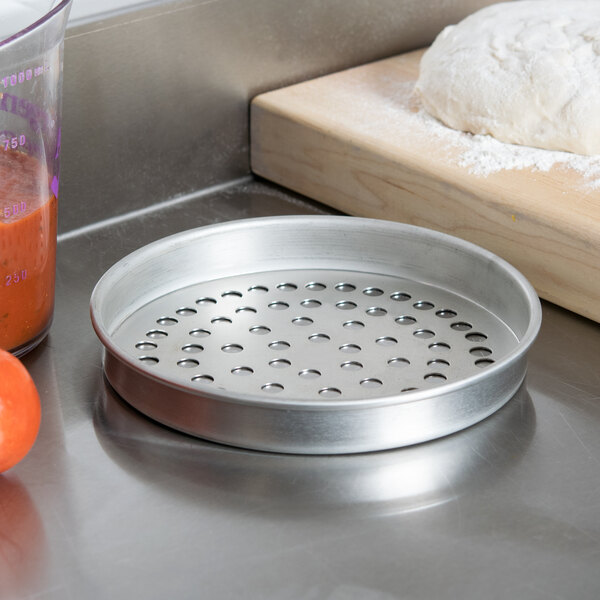 An American Metalcraft Super Perforated Pizza Pan on a table with dough and a measuring cup.