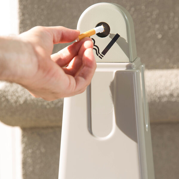 A hand holding a cigarette over a Rubbermaid beige cigarette receptacle.