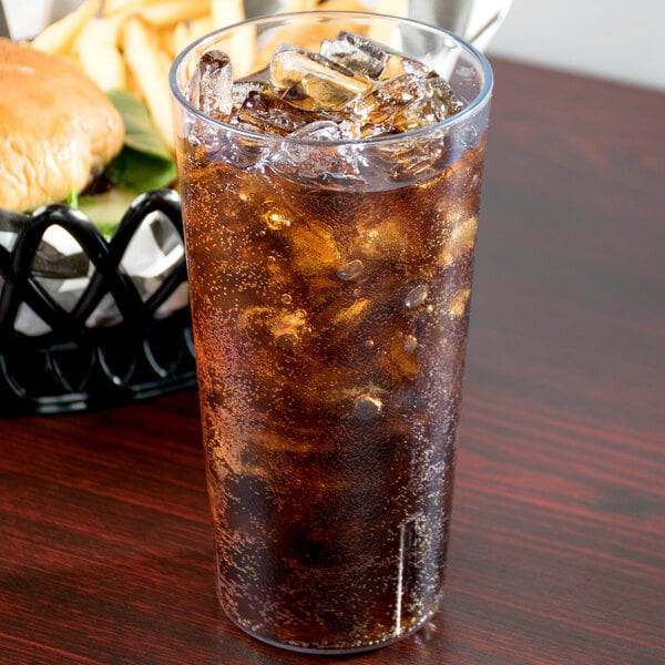 A clear plastic tumbler of soda with ice on a table with a burger in the background.