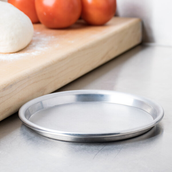 An American Metalcraft aluminum pizza pan on a counter next to tomatoes.