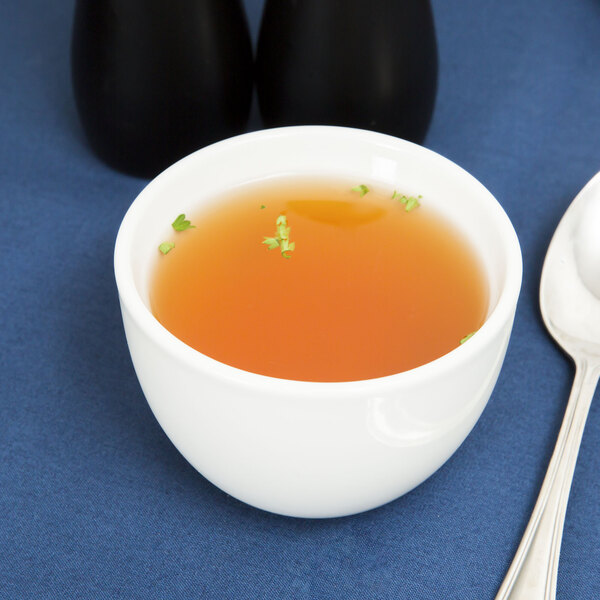 A Tuxton bright white china bouillon cup filled with soup on a blue surface with a spoon.