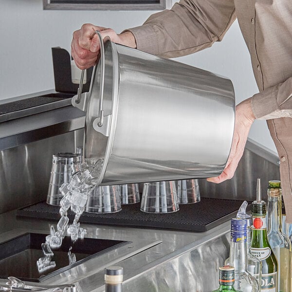 A man pouring ice into a Vollrath stainless steel dairy bucket on a counter in a cocktail bar.