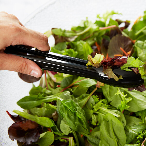 A close-up of a hand holding Carlisle black plastic utility tongs with lettuce.