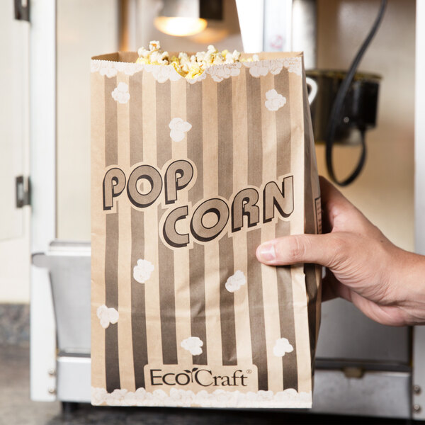 A hand holding a Bagcraft Packaging EcoCraft paper bag of popcorn.