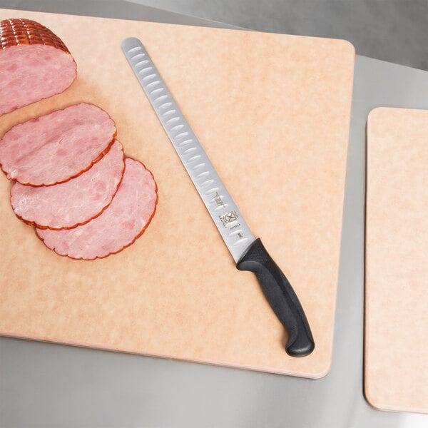 A Mercer Culinary Millennia Granton Edge Slicer Knife with a black handle next to a cutting board with sliced ham.