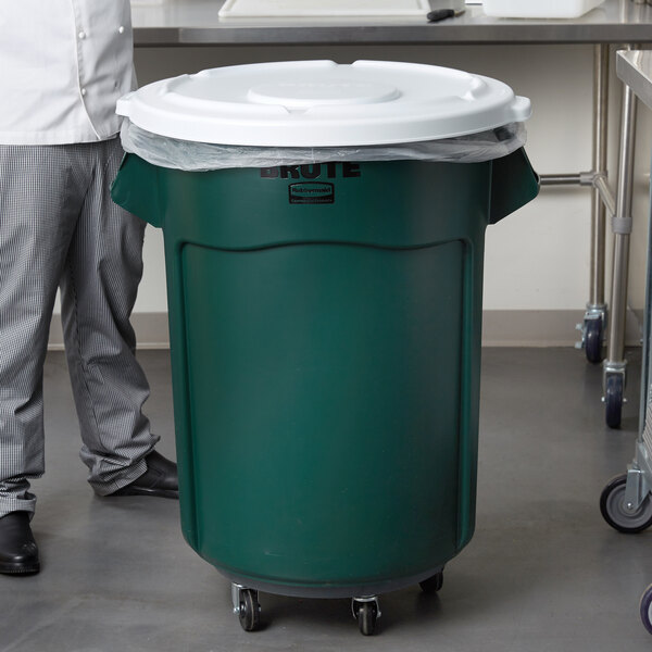 A man standing next to a green Rubbermaid BRUTE trash can with a white lid.