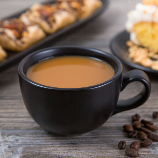 A close-up of a Libbey Driftstone Onyx Satin matte black porcelain cup filled with coffee on a table with pastries.