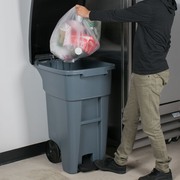 A person using a Rubbermaid wheeled step-on trash can to put a plastic bag inside.