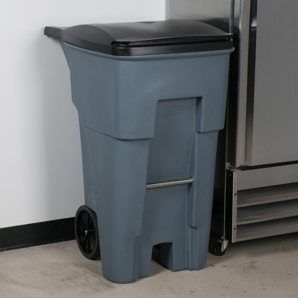 A Rubbermaid 65 gallon gray trash can with black lid next to a refrigerator.